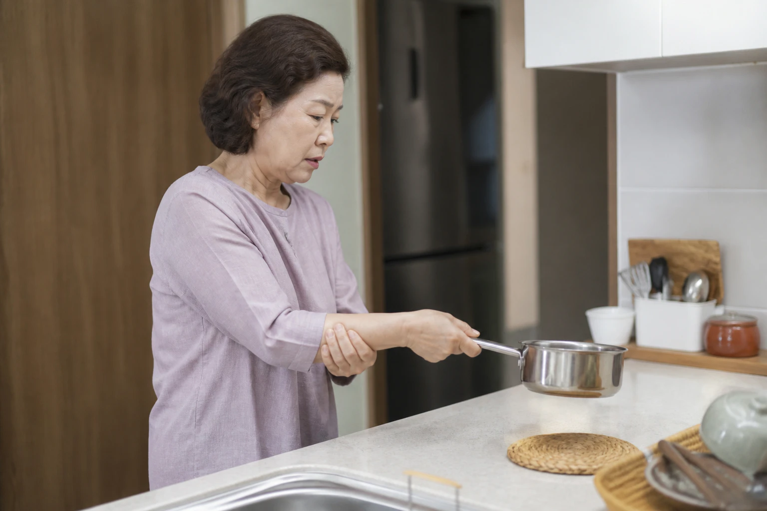 손목이 뻣뻣하고 아파 작은 냄비를 들다 다시 내려놓은 한국인 여성 한 명의 주방 장면