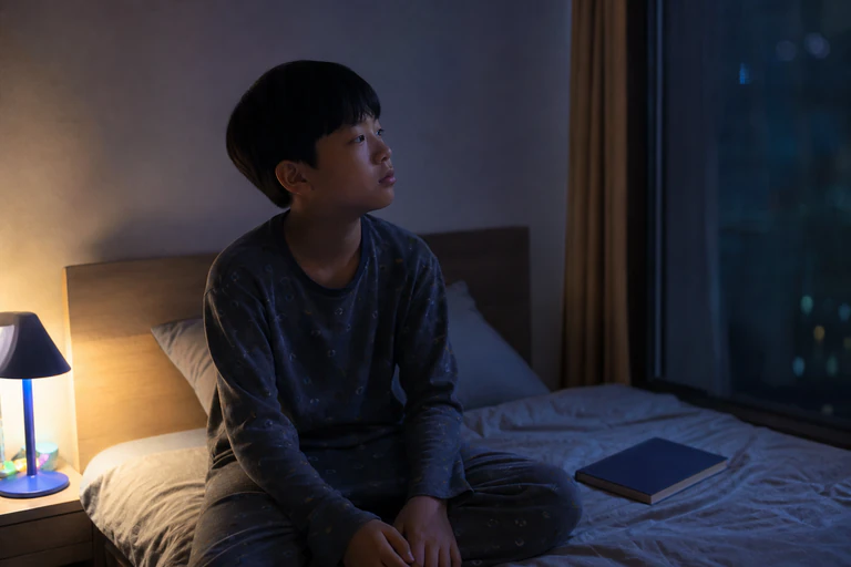 밤 침대에 앉아 잠들지 못하고 닫힌 동화책을 바라보는 한국인 남아
